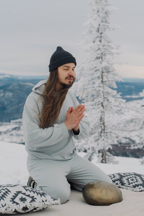 Full body of peaceful male with namaste gesture sitting on knees on snowy ground near steel tongue drum during meditation in highland