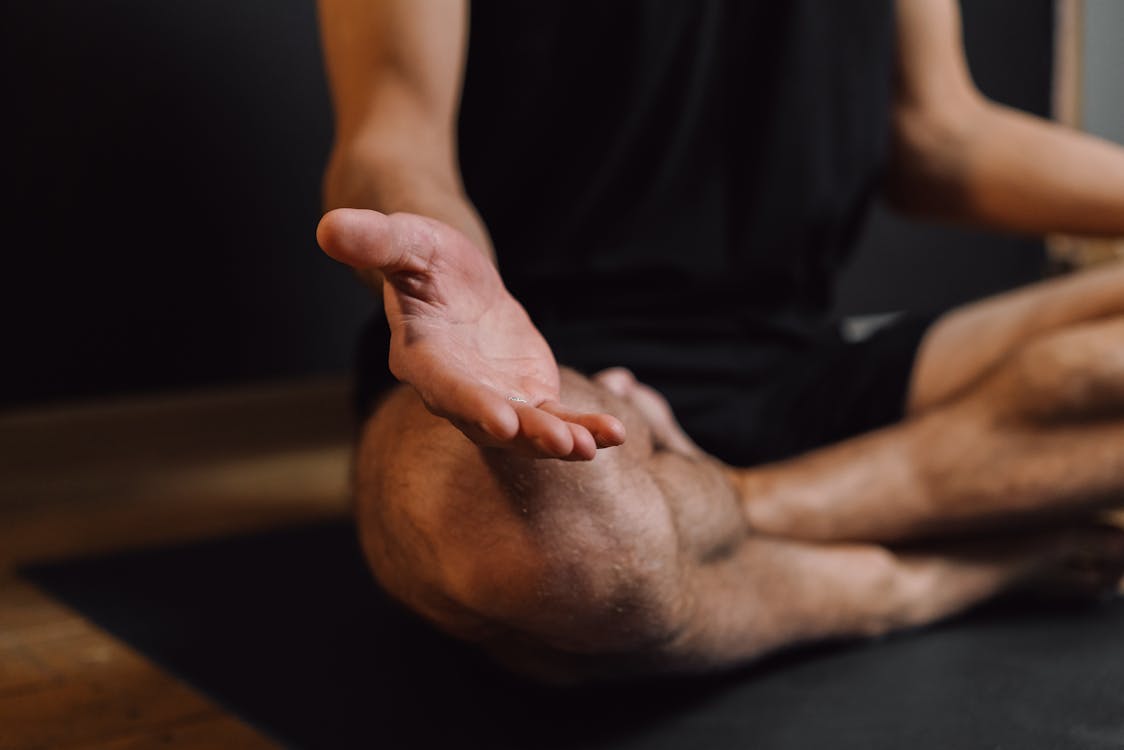 Crop unrecognizable barefoot male sitting with crossed legs on sports mat during stress relief meditation session