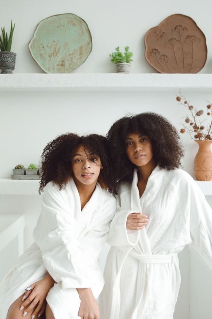 Two women in white bathrobes with afro hair posing indoors, conveying relaxation and femininity.
