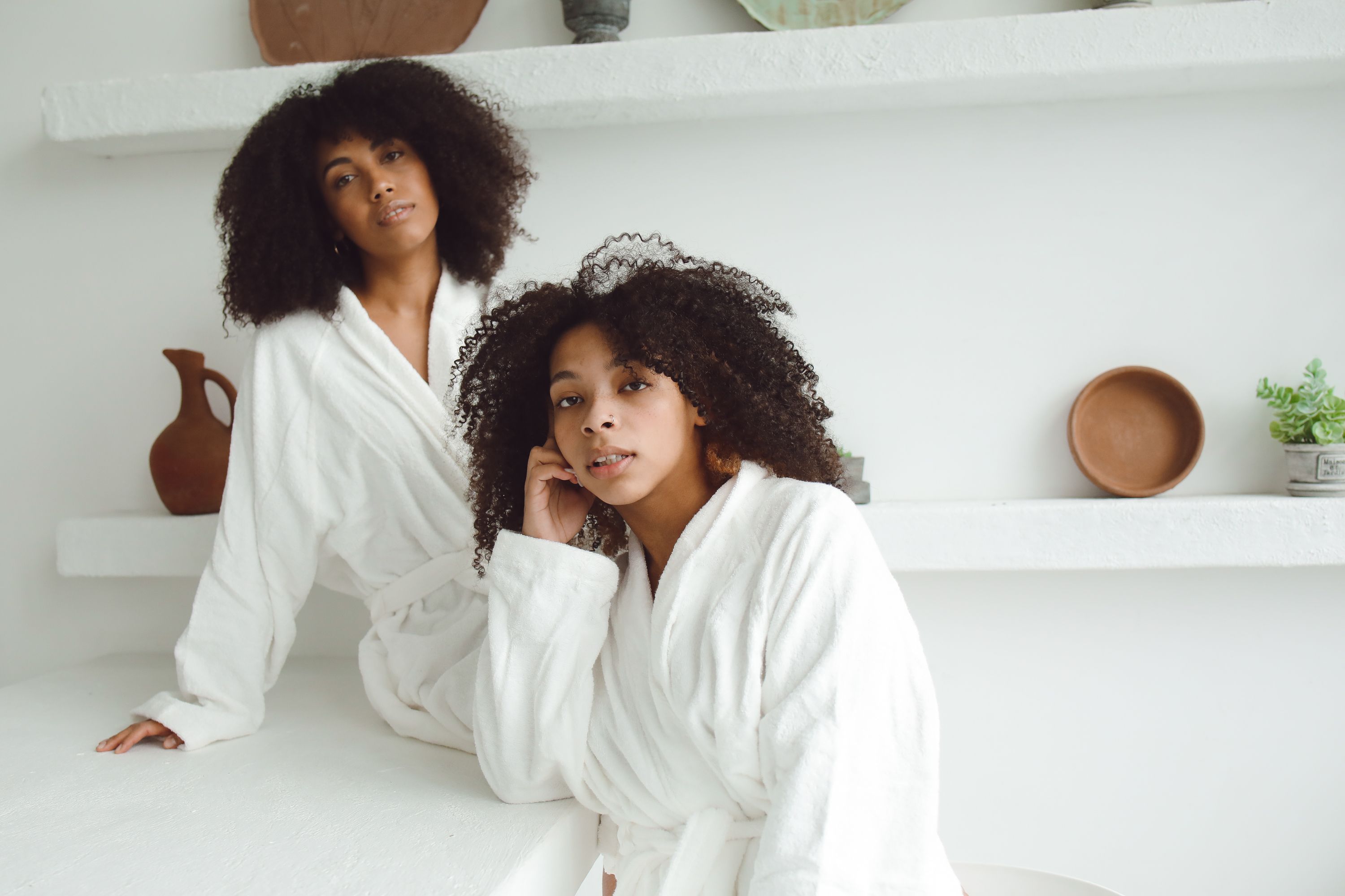 Two women in white bathrobes posing gracefully in a minimalist indoor setting.