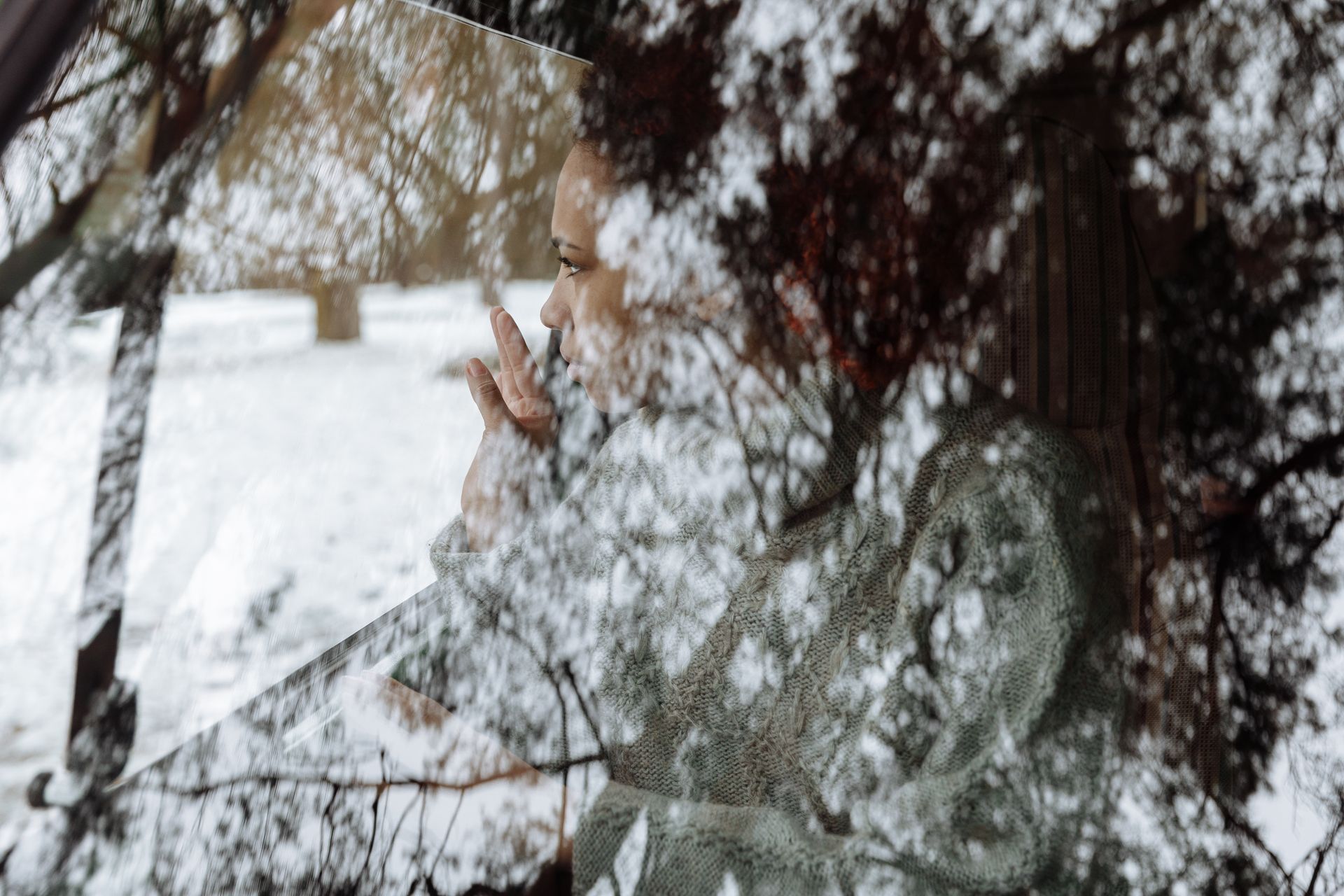 A contemplative view of a woman through a car windshield, capturing reflections of trees in winter.