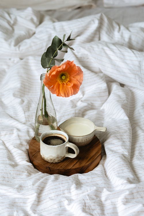 High angle of cup of aromatic coffee and milk jug placed on round wooden tray with vase of delicate fresh flower on soft bed in sunny morning