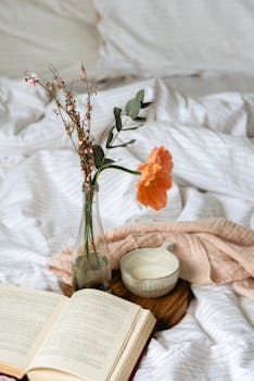 A relaxing bedside scene with a vase of flowers, open book, and a cup on a wooden board.