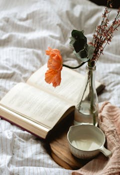 A warm and inviting setup with a book, flower, and milk on bed linen, perfect for relaxation.