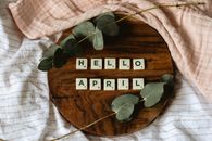 Scrabble tiles spelling 'Hello April' surrounded by eucalyptus on a wooden tray.