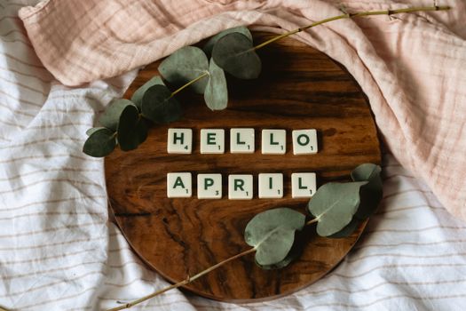 Scrabble tiles spelling 'Hello April' surrounded by eucalyptus on a wooden tray.