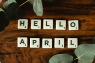 Scrabble tiles arranged to spell Hello April on a wooden surface with green leaves decor.