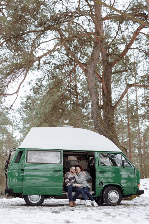 Free Couple enjoying a winter camping trip in a green camper van under snowy trees. Stock Photo