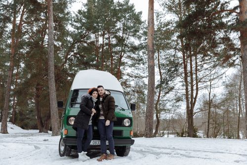 Free Couple in winter clothing embracing by a green campervan in a snowy forest setting, reflecting a cozy outdoor adventure. Stock Photo