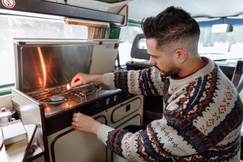 Free A man in a knitted sweater lighting a stove in a camper van kitchen, embodying cozy van life. Stock Photo