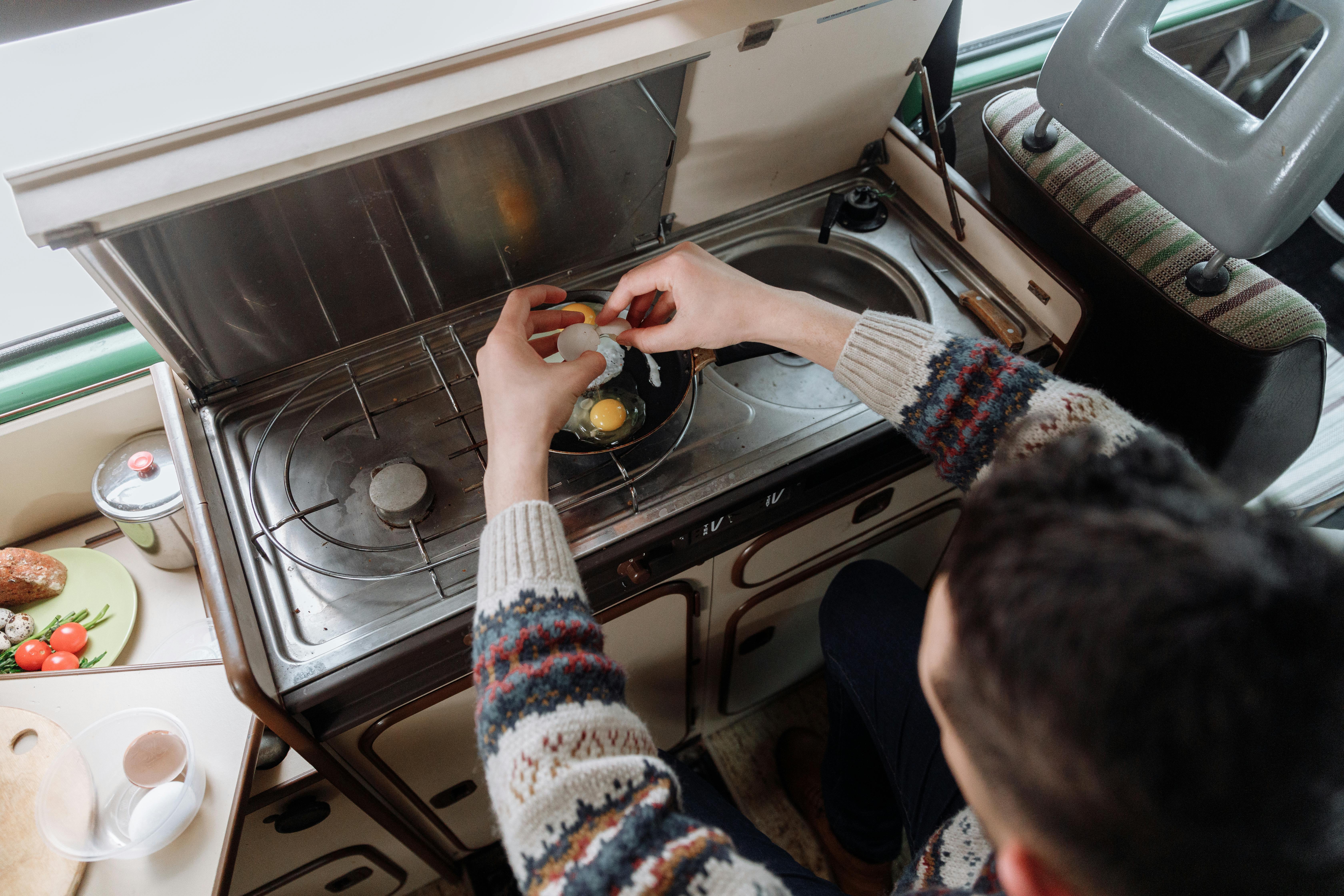 A person cracking eggs into a pan on a compact indoor stove setup. Cozy atmosphere.