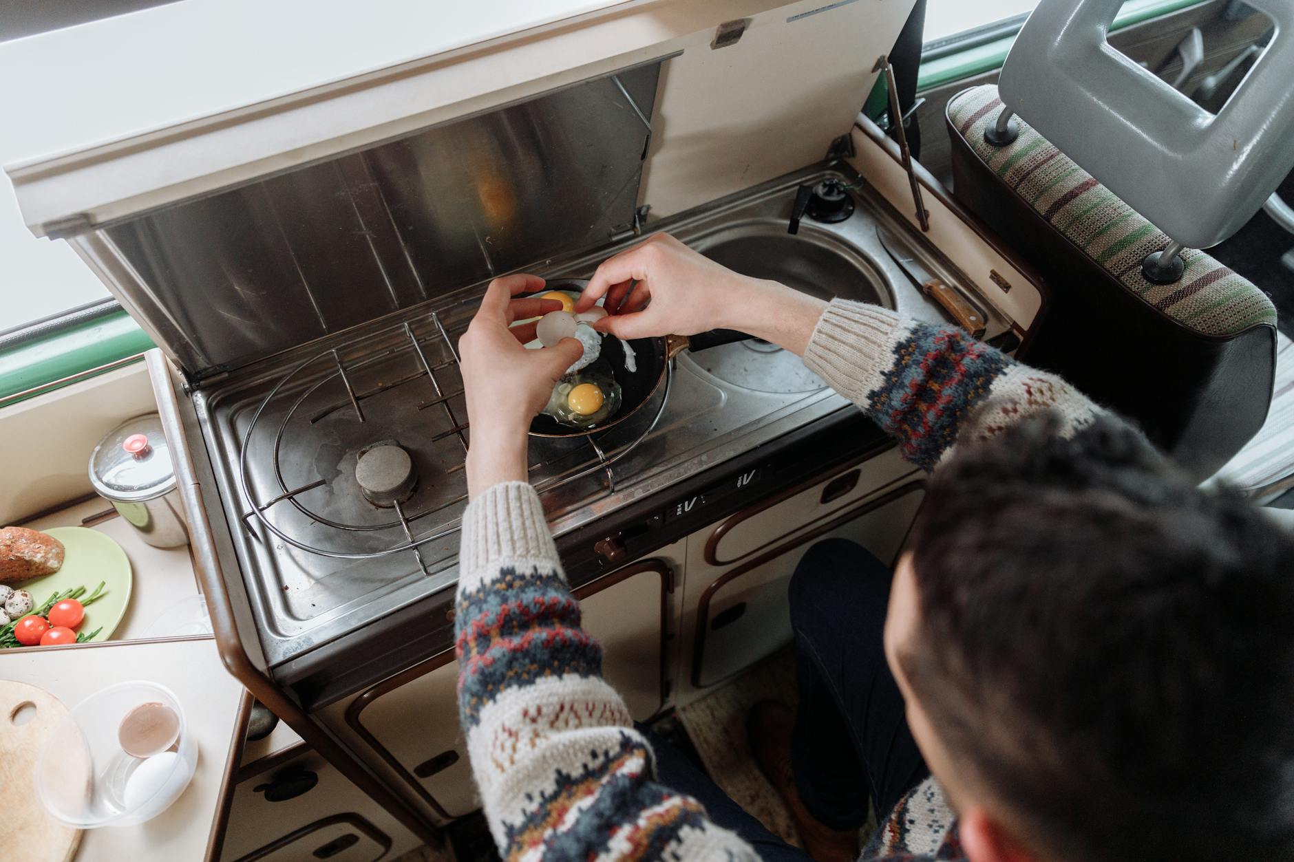 A person cracking eggs into a pan on a compact indoor stove setup. Cozy atmosphere.