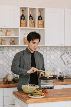 Asian man cooking healthy pancakes with fresh ingredients in a stylish home kitchen.
