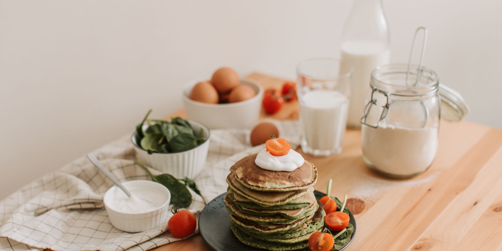Delicious stack of pancakes with fresh spinach, tomatoes, eggs, milk, and yogurt on a wooden table.
