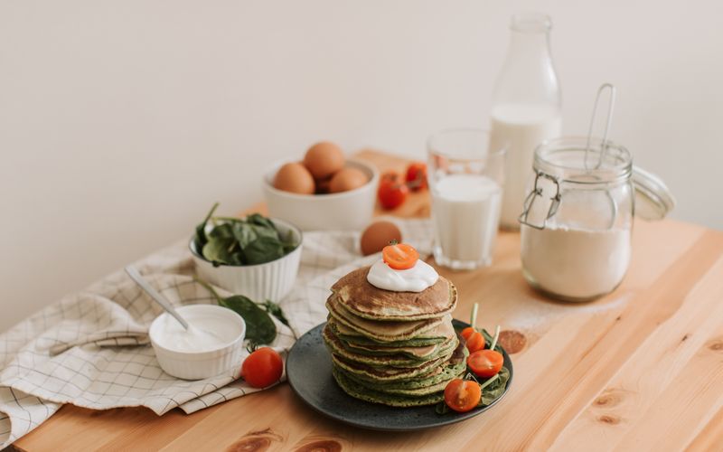 Delicious stack of pancakes with fresh spinach, tomatoes, eggs, milk, and yogurt on a wooden table.