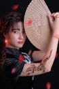 Close-Up Shot of a Pretty Woman in Black Traditional Dress Holding a Hand Fan while Looking at Camera