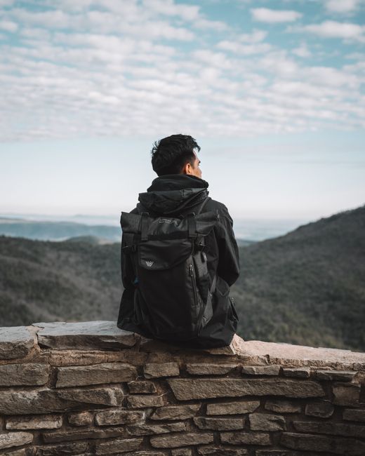 A man sits on a stone ledge with a backpack, enjoying a scenic mountain view under a cloudy sky.