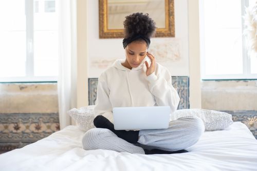 Free A woman in a hoodie works on a laptop, sitting cross-legged in a well-lit bedroom. Stock Photo