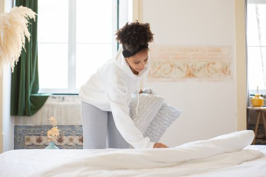 An African American woman arranged pillows while making a bed in a bright and cozy room.