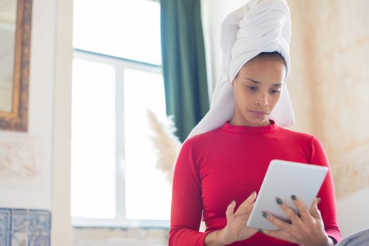 African American woman with a towel wrapped around her head, using a tablet indoors.