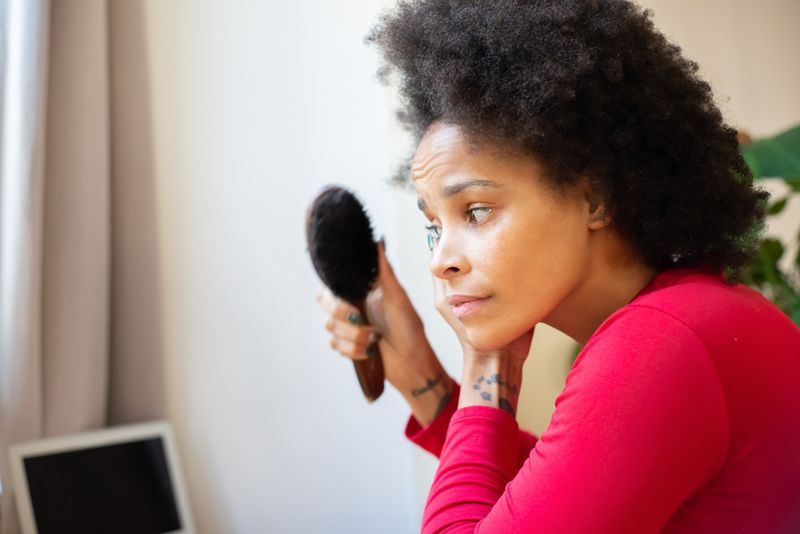 A Black woman in a red top holds a hairbrush loaded with shed hair, her brow furrowed.