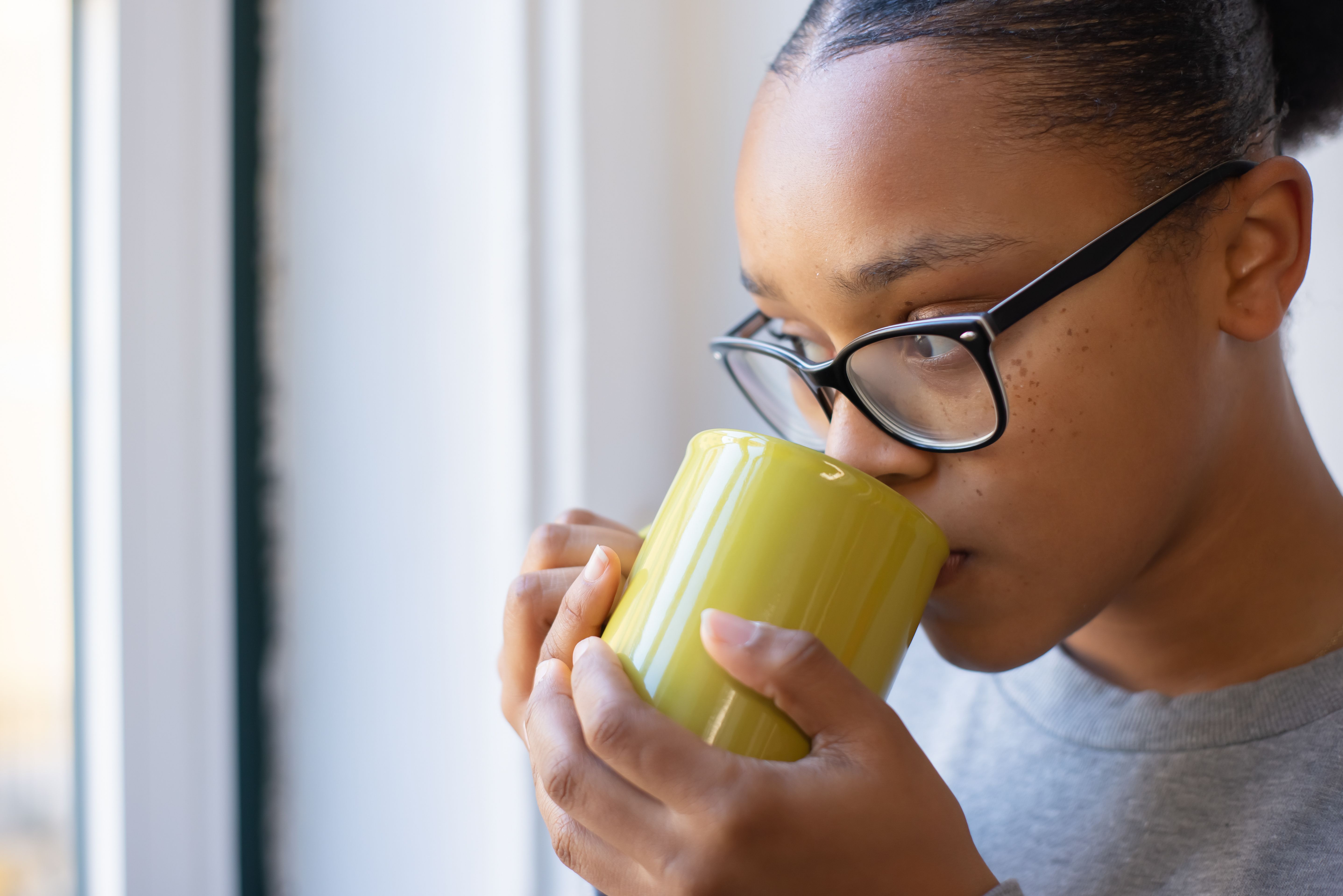 Close-up of a woman with eyeglasses sipping a hot drink near a window.