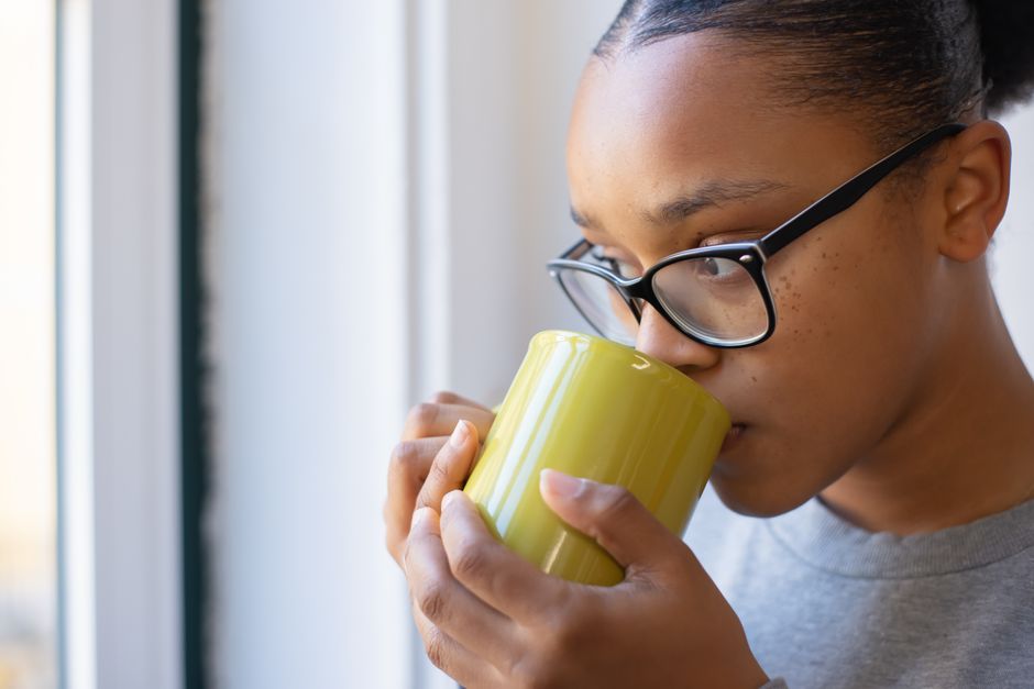 Close-up of a woman with eyeglasses sipping a hot drink near a window.