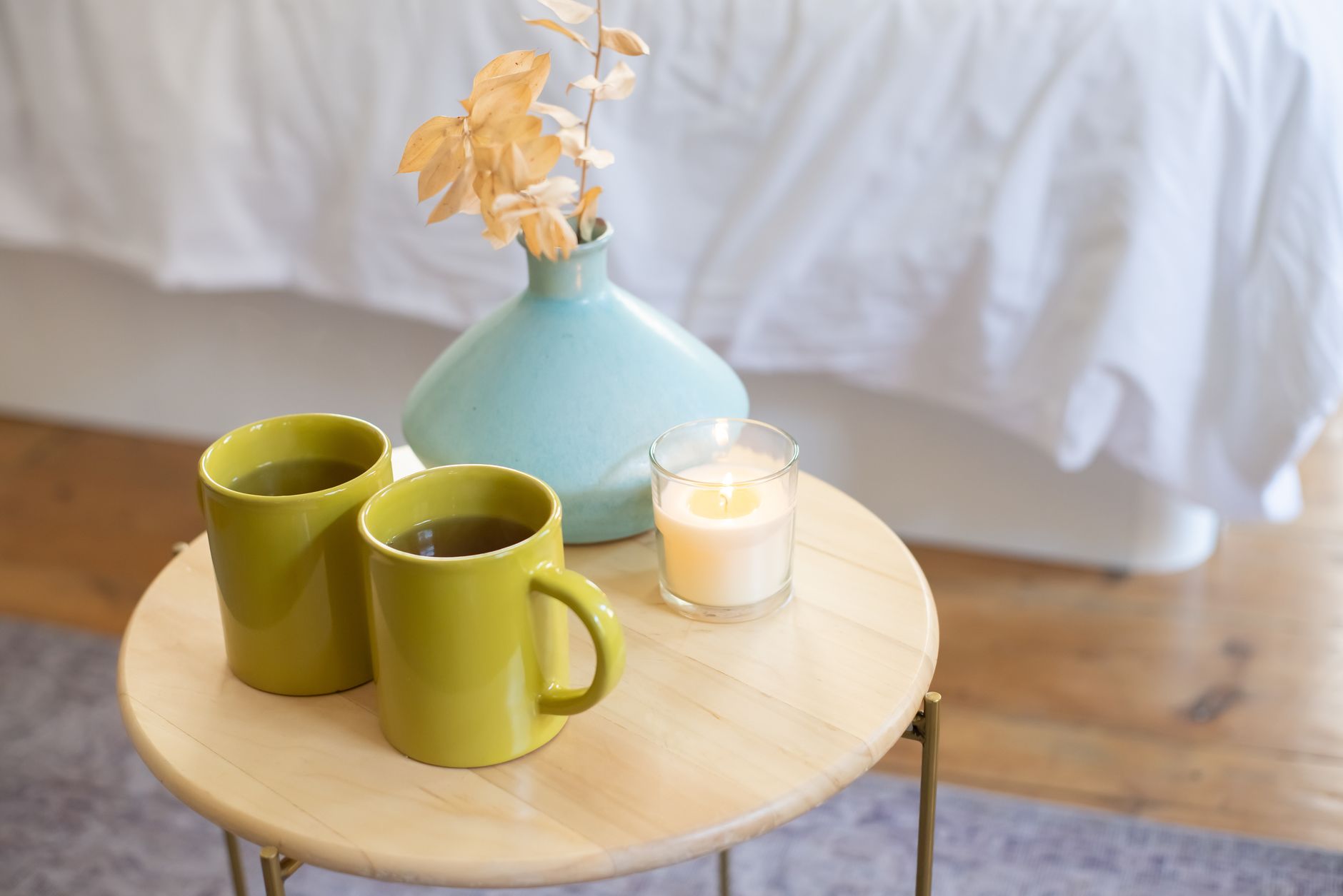 A cozy indoor scene with green mugs, lit candle, and vase on a wooden table.