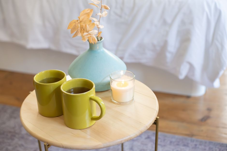 A cozy indoor scene with green mugs, lit candle, and vase on a wooden table.