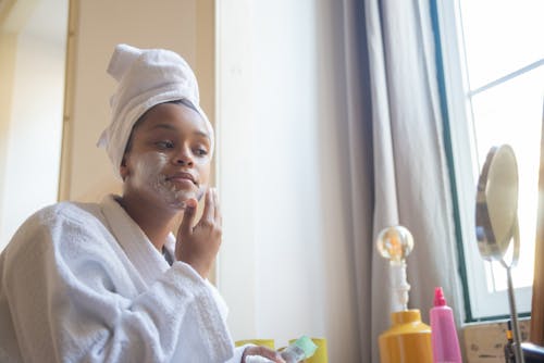 A Woman in White Bathrobe Applying a Cream on Her Face while Looking at the Mirror