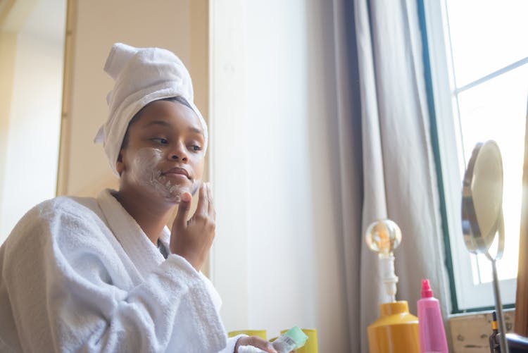 A Woman In White Bathrobe Applying A Cream On Her Face While Looking At The Mirror