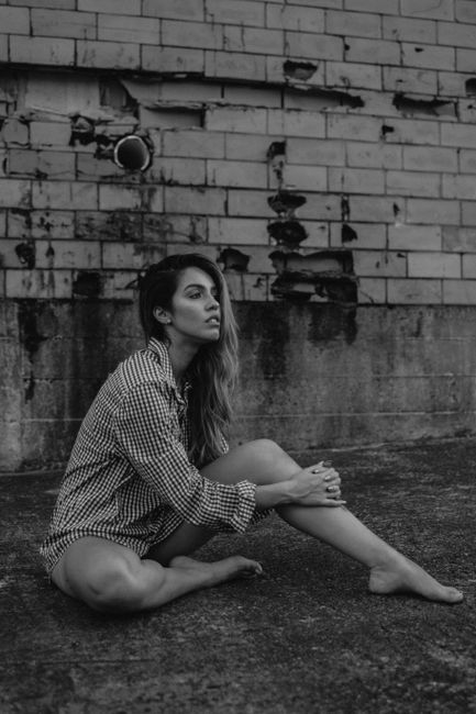 Artistic black and white portrait of a woman sitting against an aged brick wall outdoors.