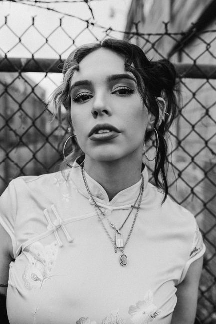 Striking monochrome portrait of a young woman with hoop earrings and silver necklaces against a chain-link fence.