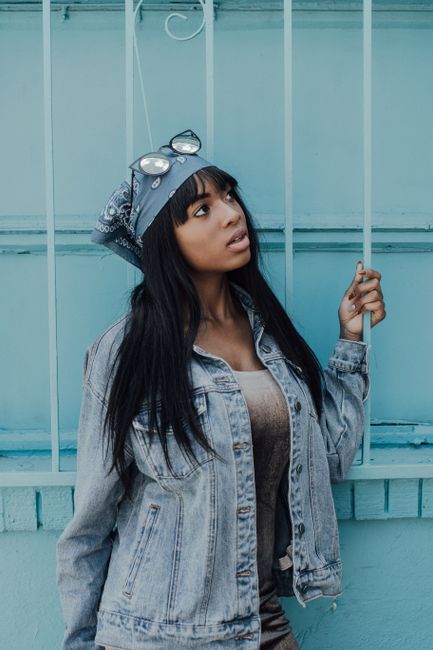 Stylish black woman in denim jacket posing against blue wall holding metal bars.