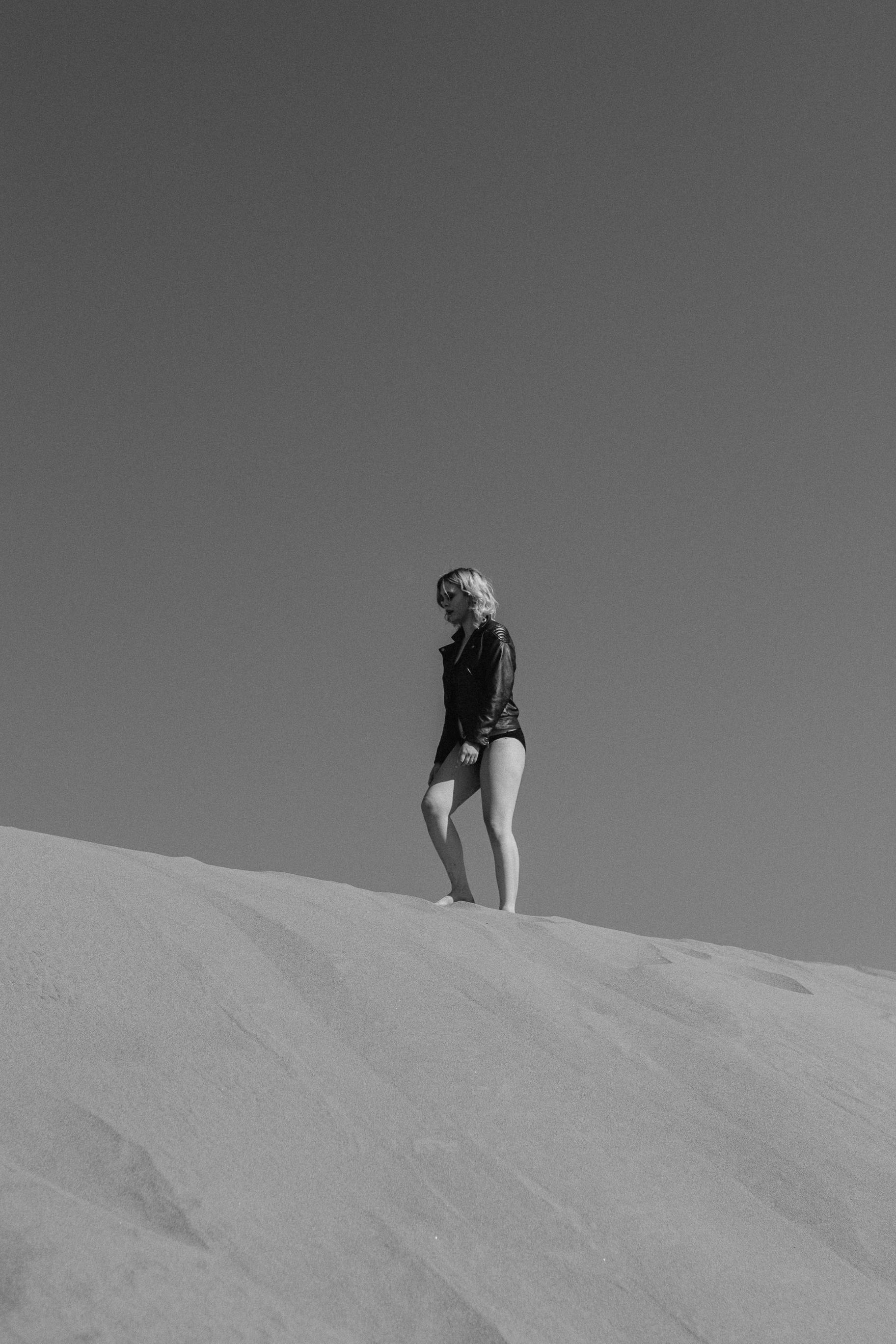 Black and white image of a woman walking on sand dunes under a clear sky.