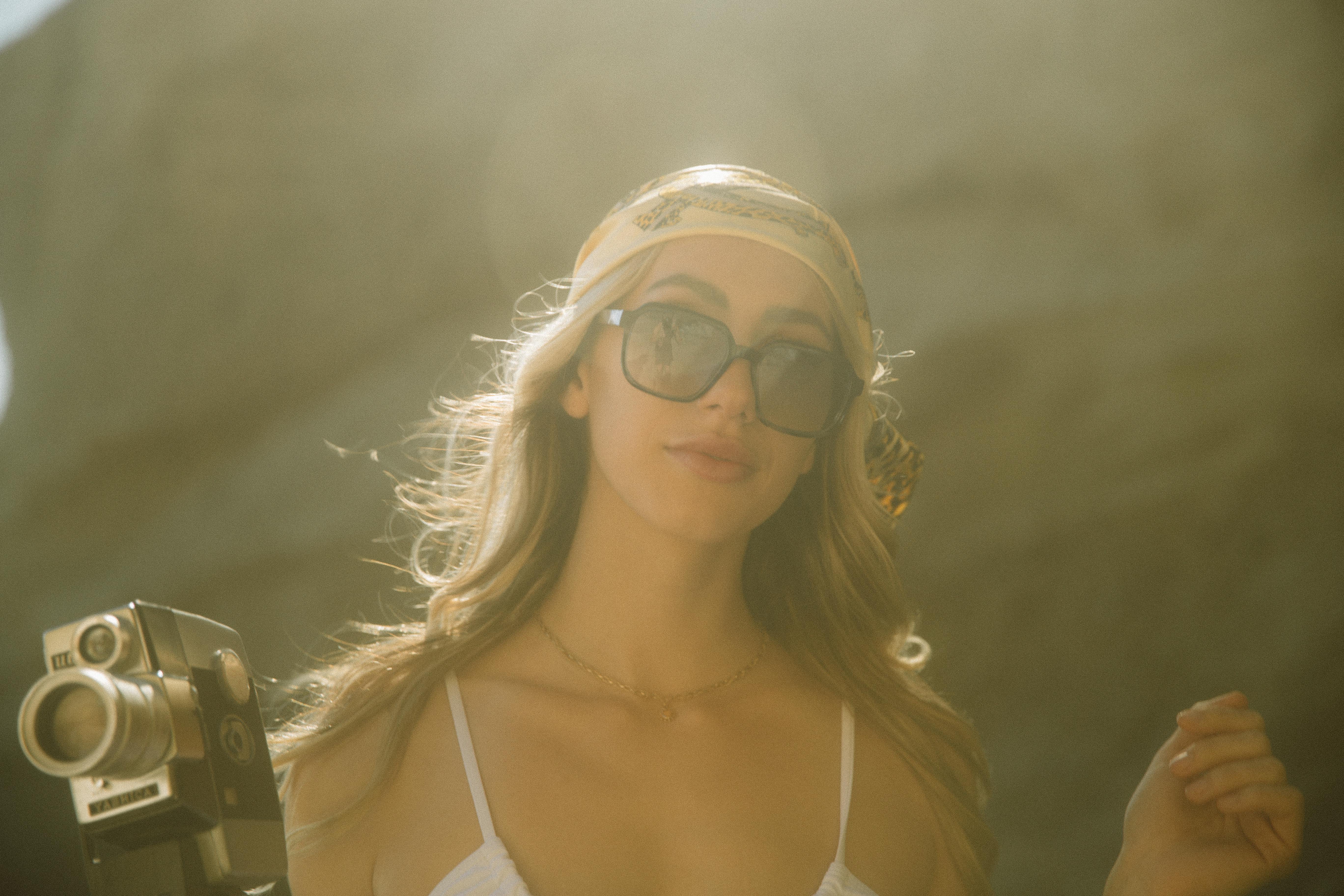 Stylish woman in sunglasses holding a vintage camera outdoors in sunlight.