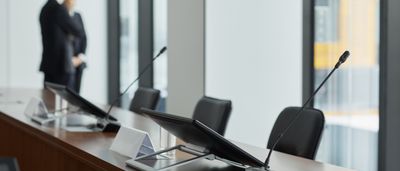 A modern corporate boardroom table with microphones and monitors, with two executives standing out of focus in the background.