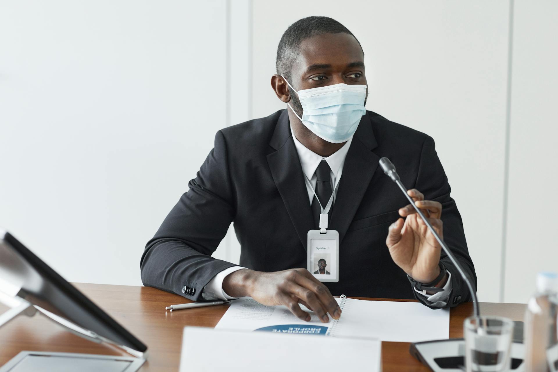 An adult male wearing a suit and face mask speaks at a conference table with a microphone.