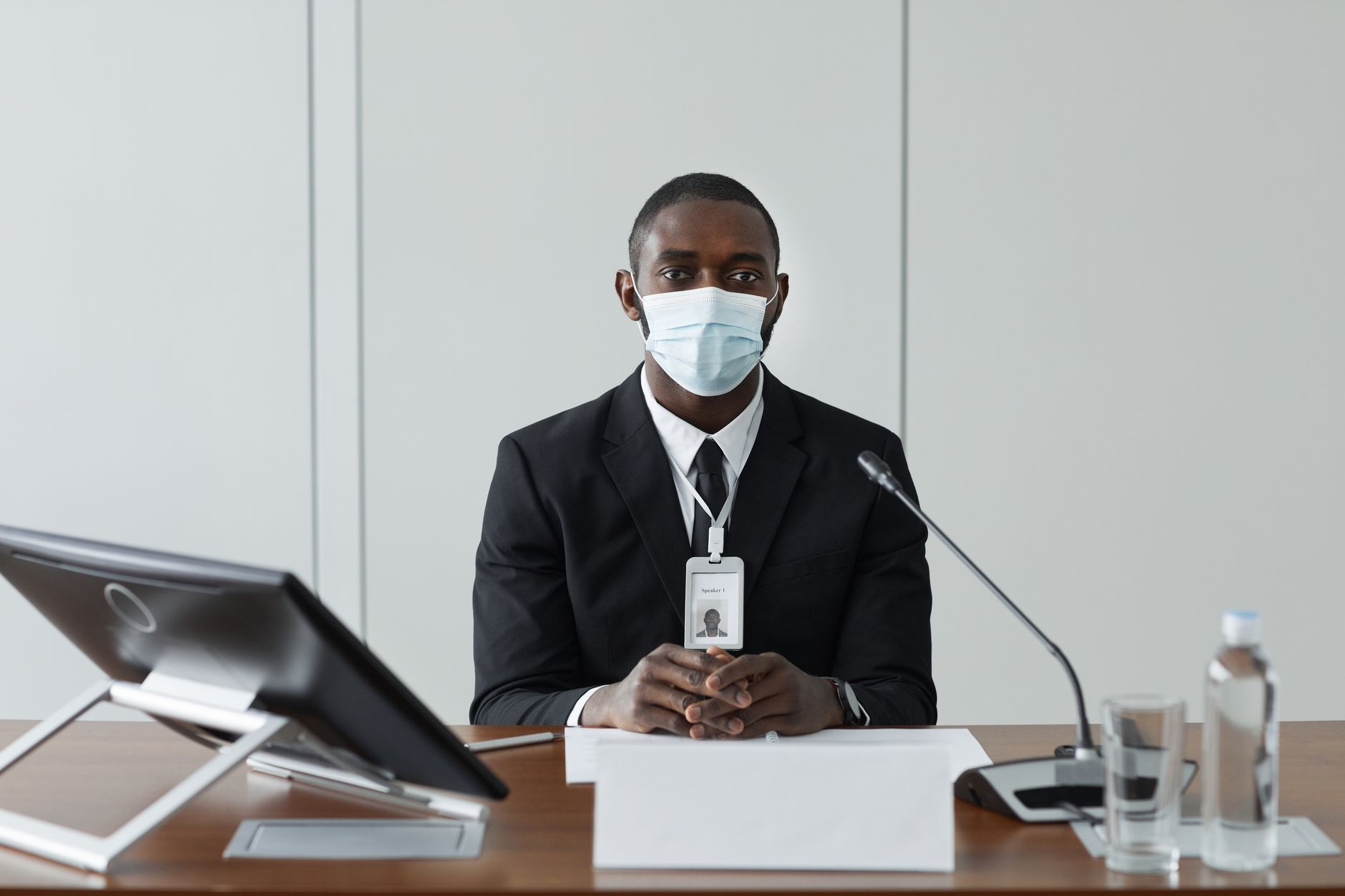 Professional African American man wearing a mask and suit, addressing a meeting at a conference table.