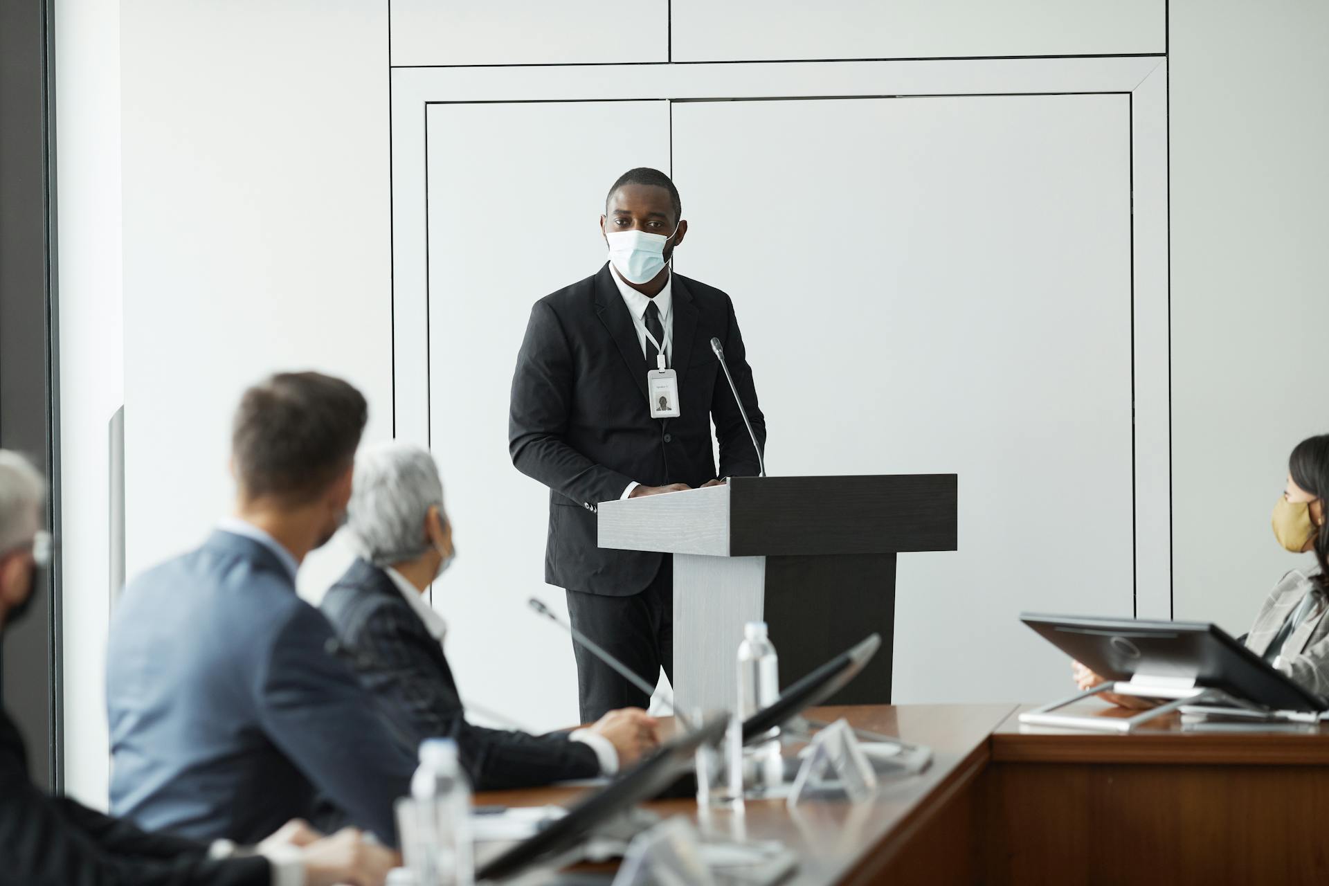 Professional speaker presenting at a business conference with colleagues seated around a boardroom table.