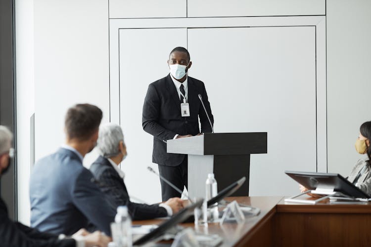 Man In A Black Suit Wearing A Face Mask Standing On A Podium