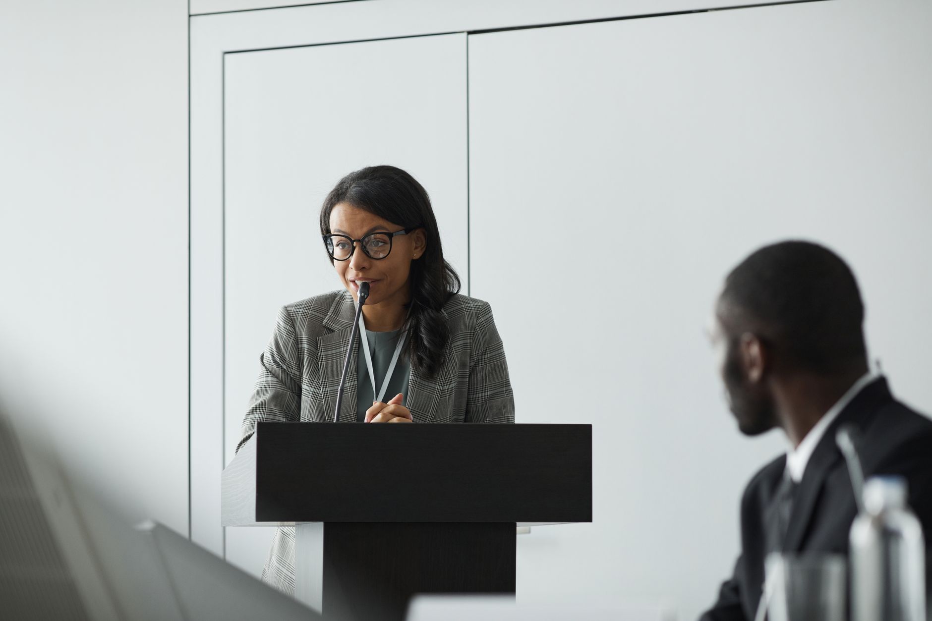 African American businesswoman delivering a speech at a conference podium with a colleague listening.