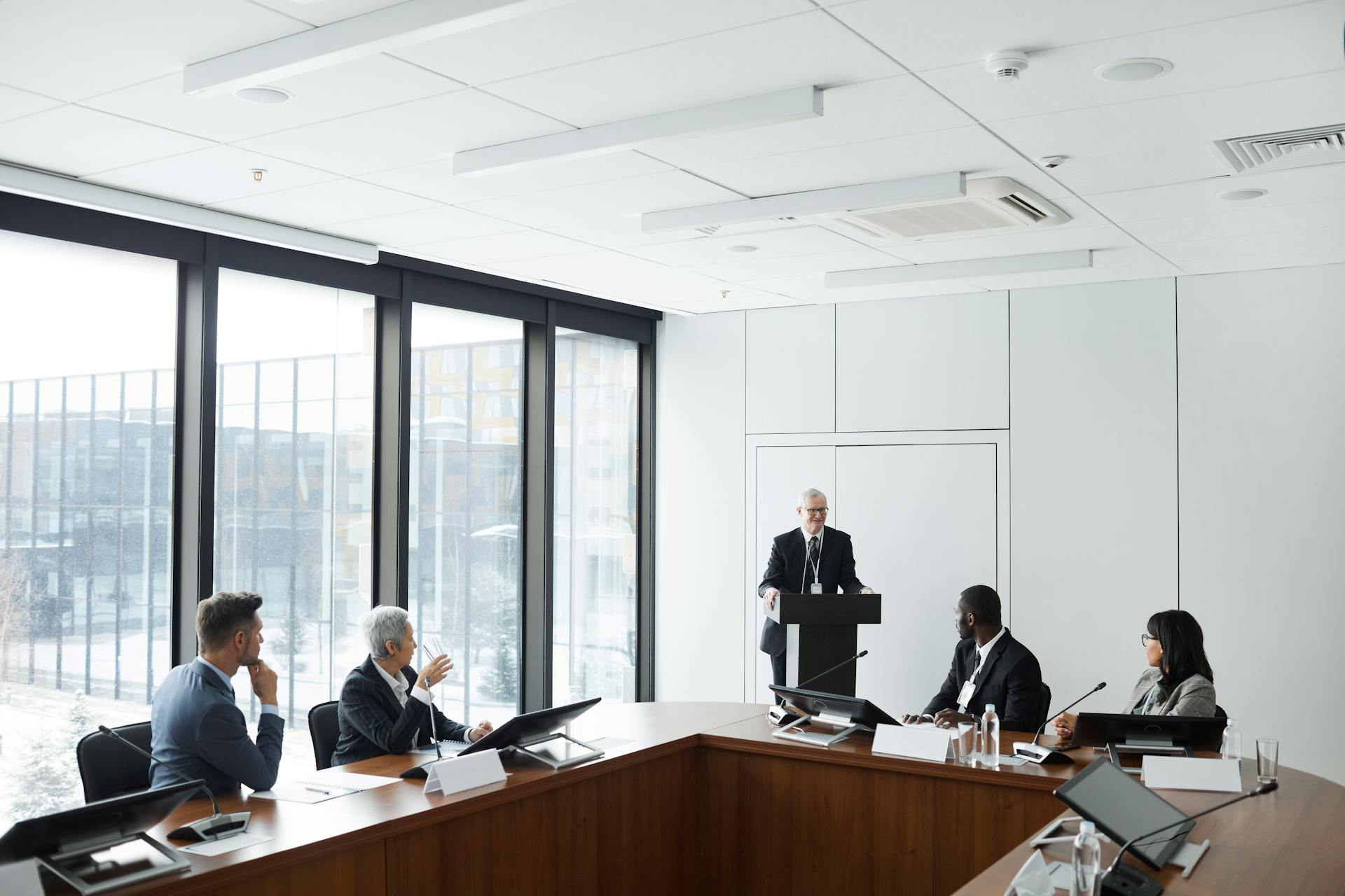 Corporate meeting with diverse professionals listening to a speaker at a podium in a modern conference room.