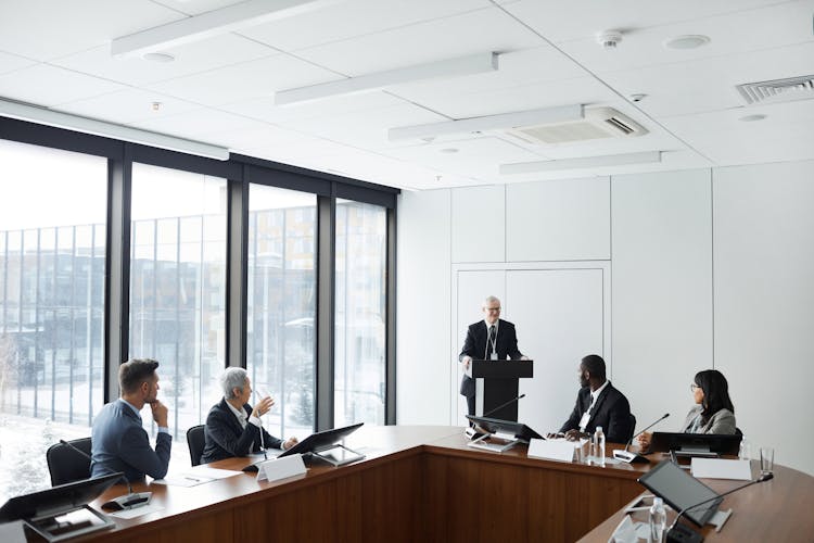 A Group Of People Sitting Near The Wooden Table While Listening To The Speaker