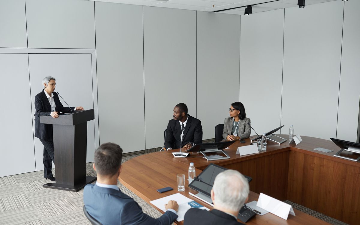 Diverse group of professionals in a formal meeting setting with a speaker at the podium.