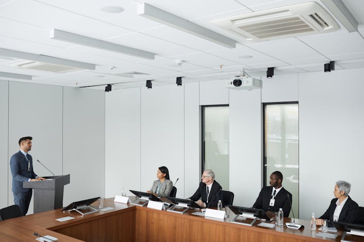 A Group Of People Sitting Near The Wooden Table While Listening To The Speaker