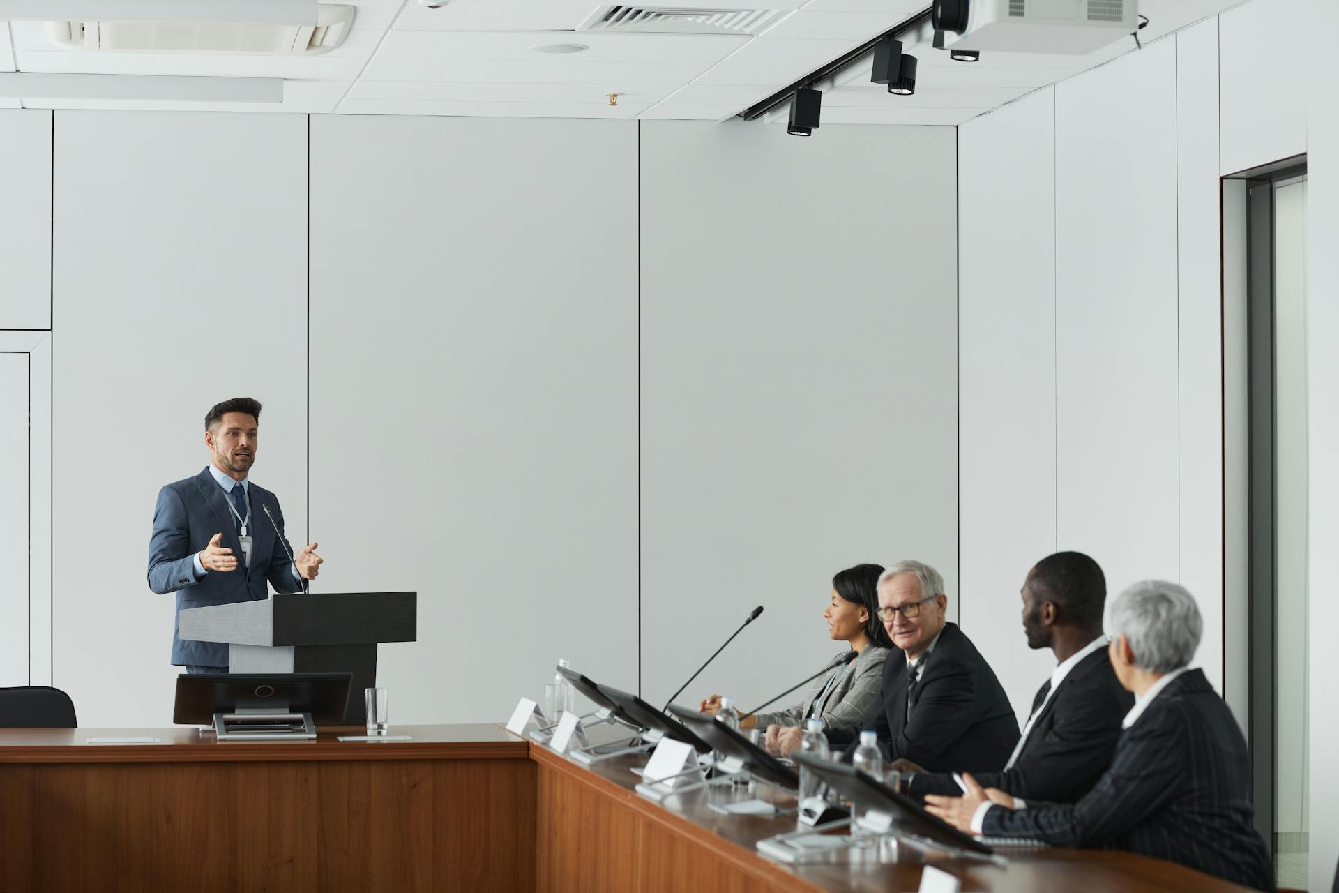 Formal business meeting with a speaker at the podium addressing a diverse panel in a conference room.