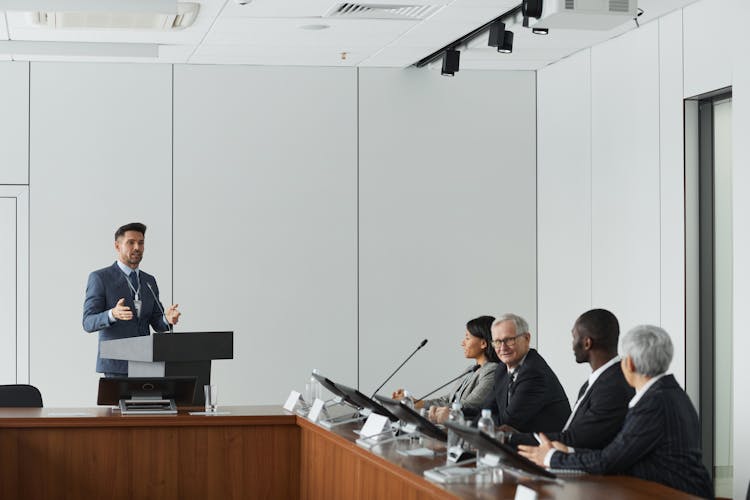 A Group Of People Sitting Near The Wooden Table While Listening To The Speaker