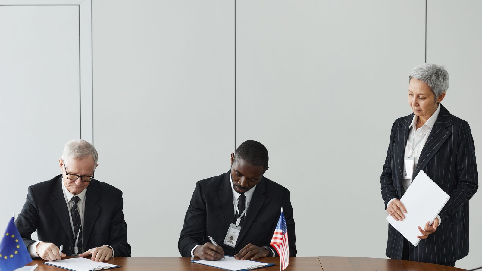 Executives signing international agreement with EU and US flags displayed on a wooden table.