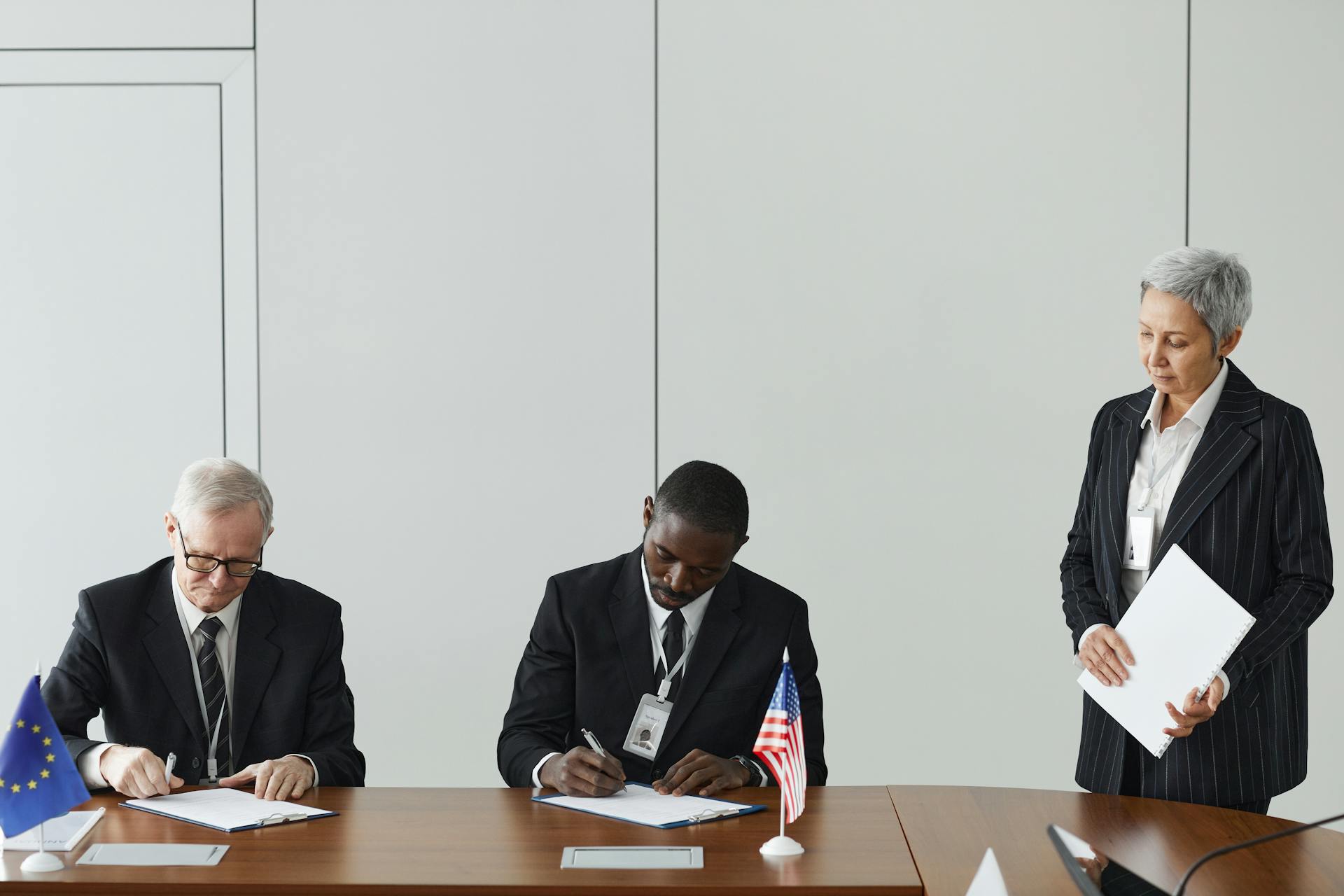 Executives signing international agreement with EU and US flags displayed on a wooden table.